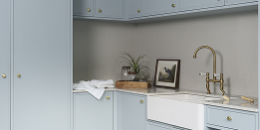 Rectangle 1 (15) A laundry room with light blue cabinets, a farmhouse sink, brass faucet, black-and-white checkered tile floor, small wooden stool, potted plant, and framed artwork on the countertop.