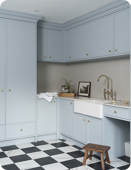 A laundry room with light blue cabinets, a farmhouse sink, brass faucet, black-and-white checkered tile floor, small wooden stool, potted plant, and framed artwork on the countertop.