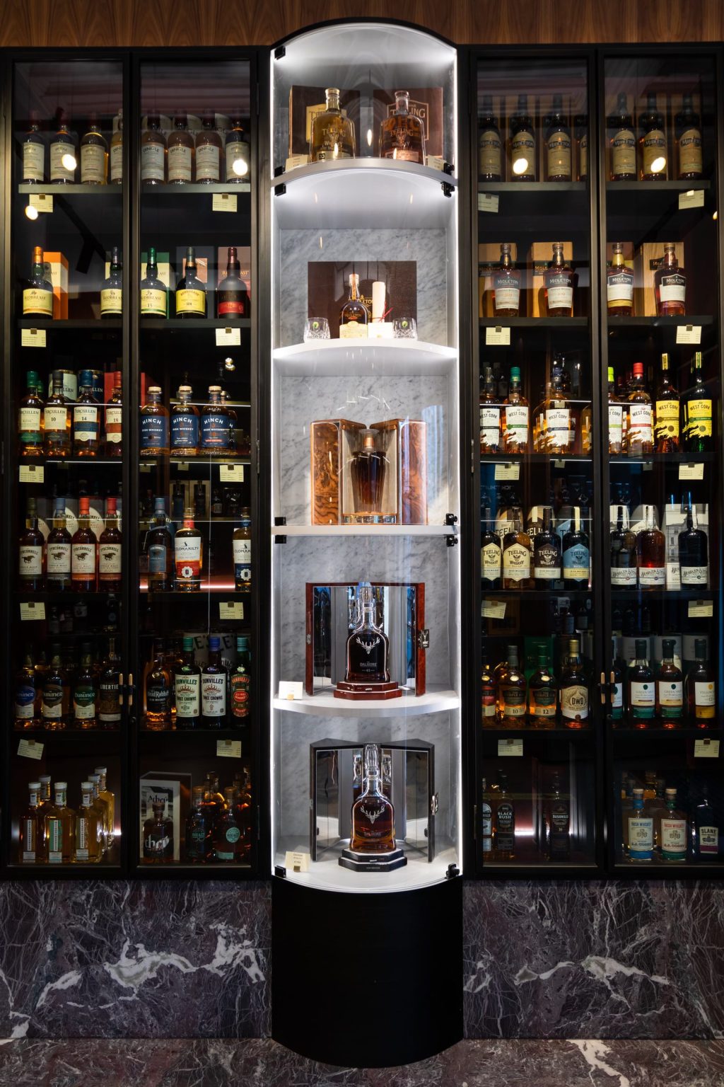 Black-framed glazed whiskey cabinets either side of a lit, curved display column on a marble plinth, in the James Fox shop on Grafton Street, Dublin