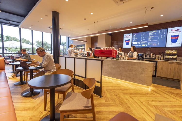 A modern café with people sitting at tables using laptops and phones. Baristas stand behind the counter with a red espresso machine. Large menu boards hang on the wall, and floor-to-ceiling windows let in natural light.