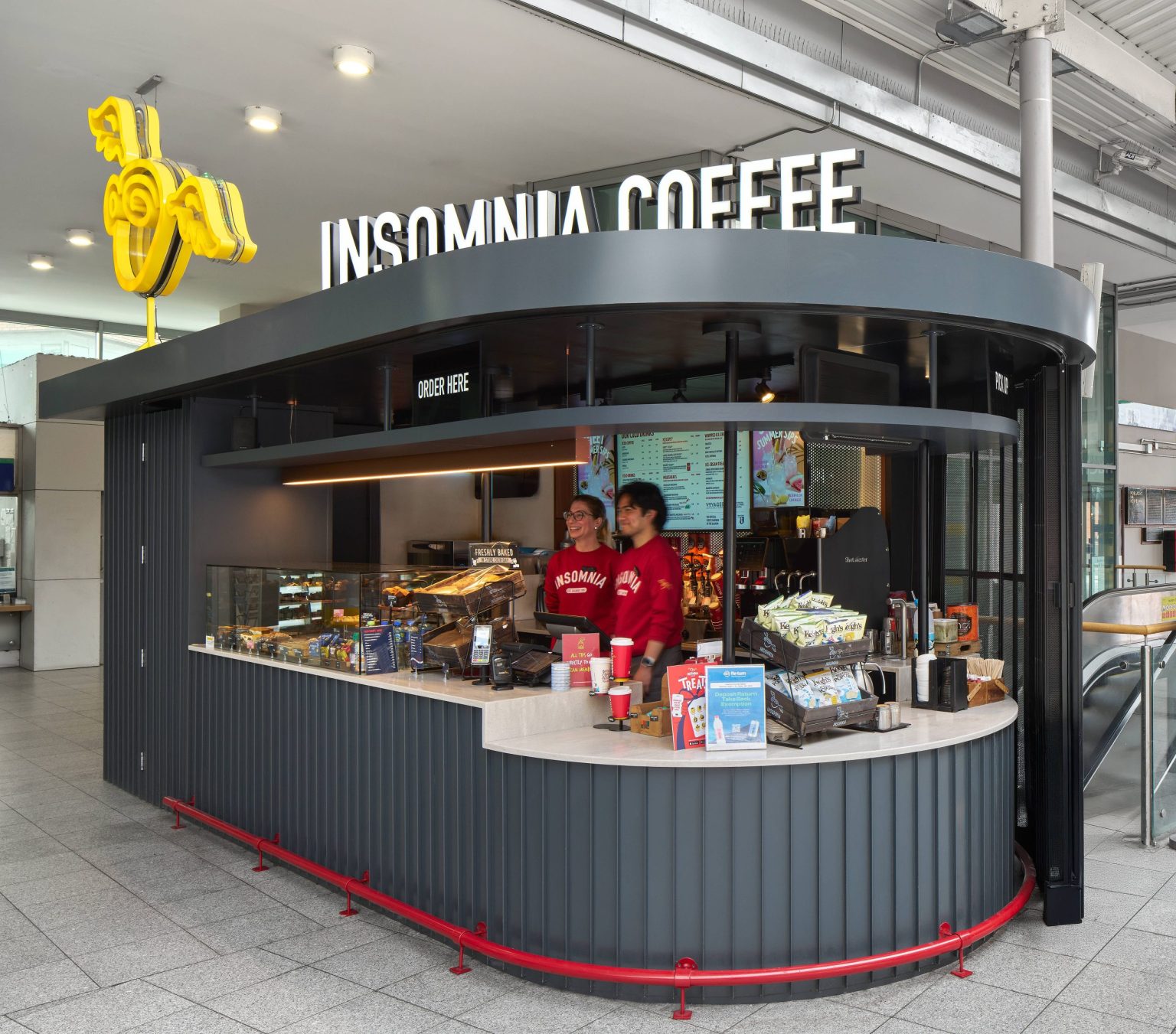 Insomnia Coffee kiosk at Connolly Station with a curved charcoal fluted counter, a red foot rail and two staff serving