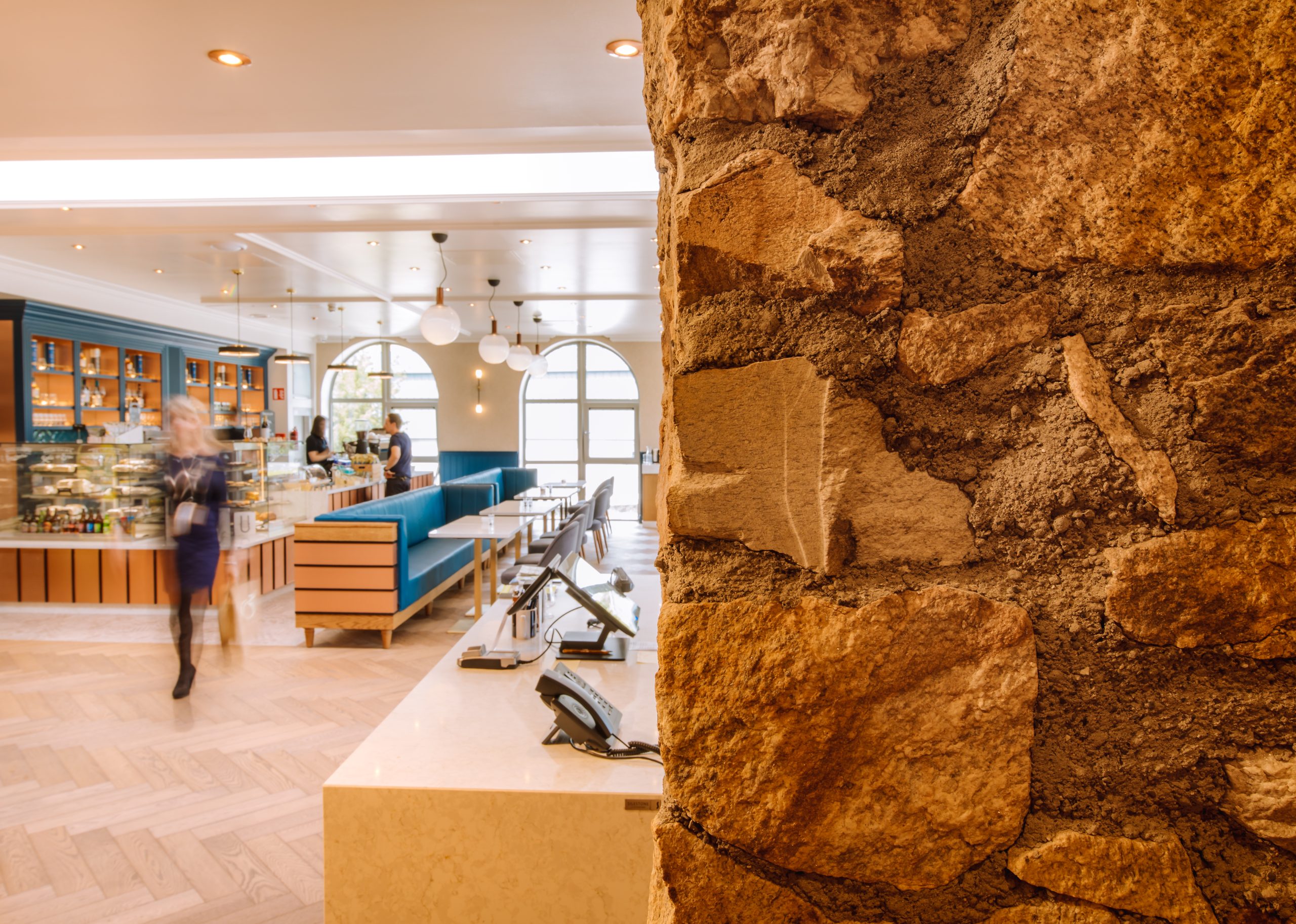 Stylish cafe interior with stone wall, herringbone floor, blue booths, and arched windows.