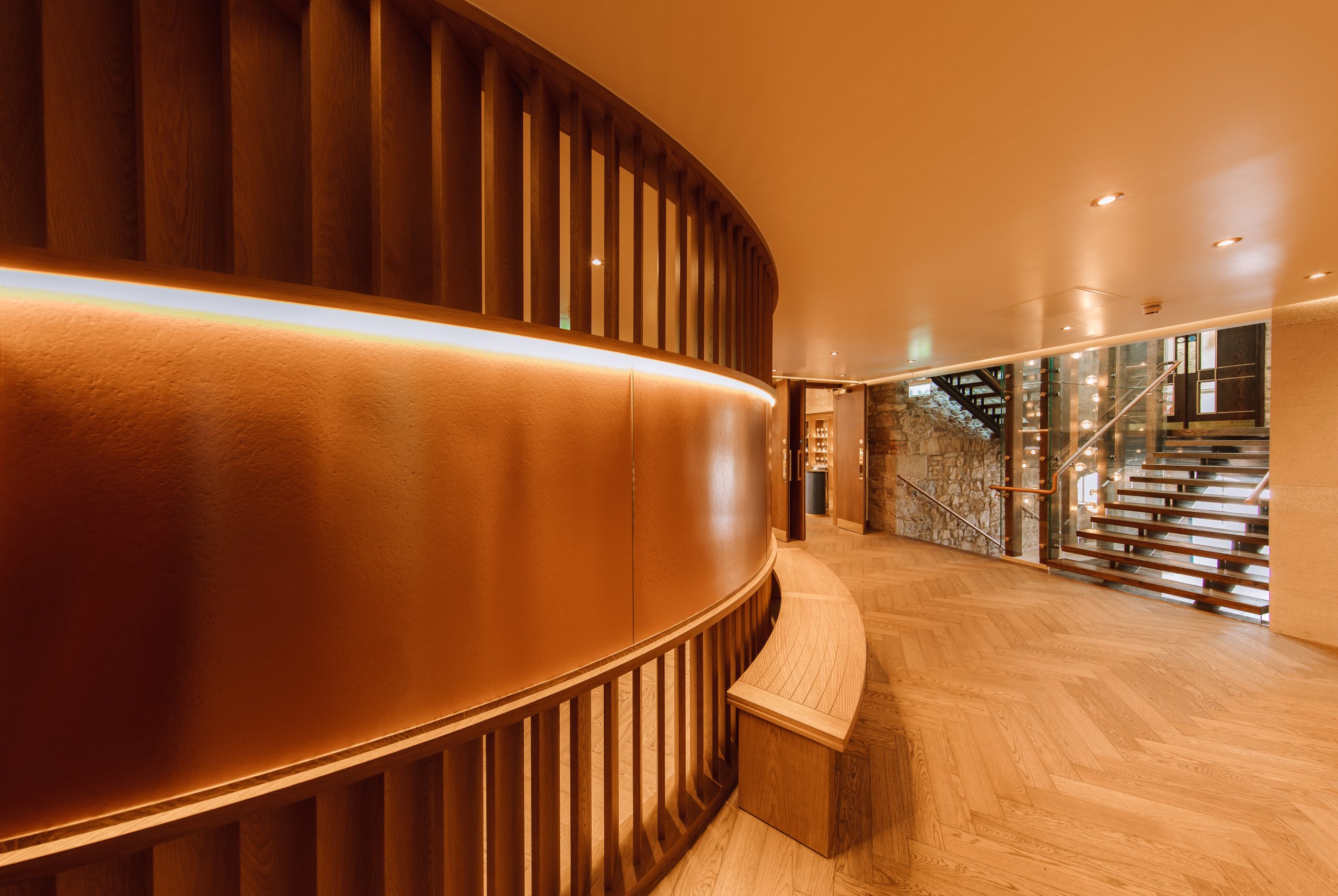 Modern hallway with curved wood wall, herringbone floor, and floating staircase.