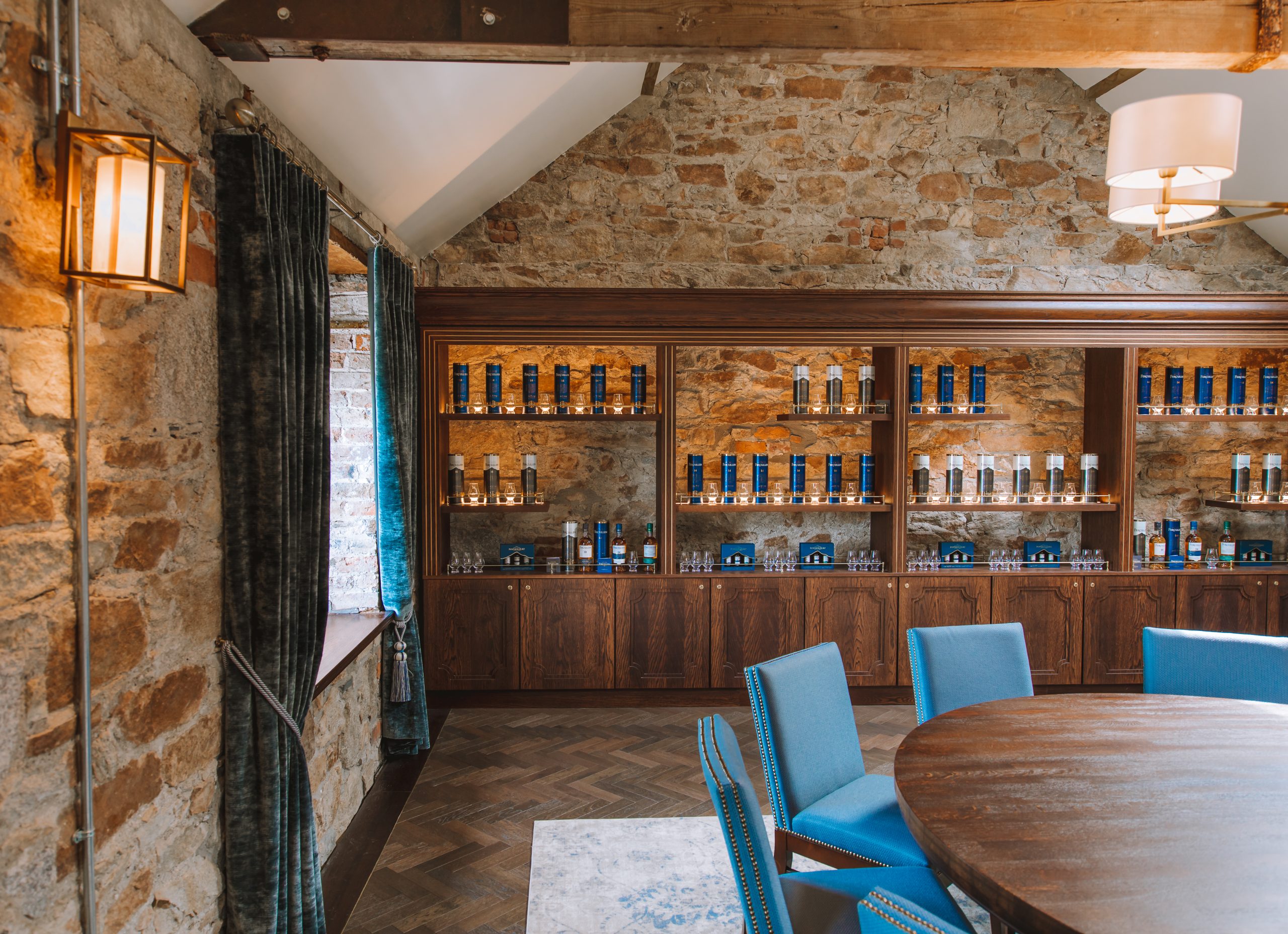 Elegant tasting room with stone walls, wooden shelves stocked with blue bottles, and blue chairs around a table.