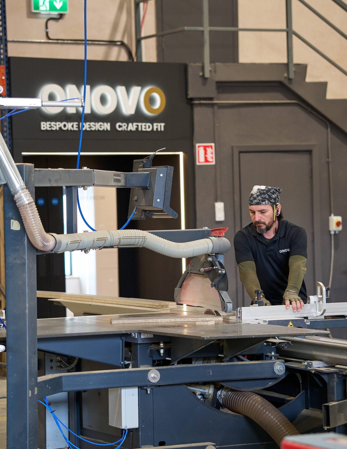 A joiner cutting a panel on a sliding table saw in the ONOVO workshop, with the lit ONOVO Bespoke Design, Crafted Fit sign behind him